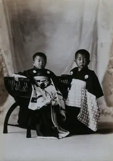 Two boys in a photographic studio, one seated on a chair, in front of a curtained backdrop, c1900. Creator: Unknown