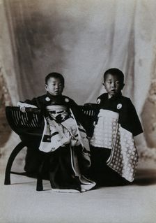 Two boys in a photographic studio, one seated on a chair, in front of a curtained backdrop, c1900. Creator: Unknown