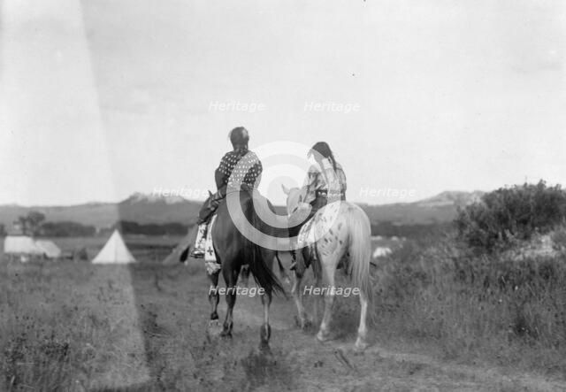 Two daughters of a chief on horseback, riding away from camera toward tents in background, c1907. Creator: Edward Sheriff Curtis.