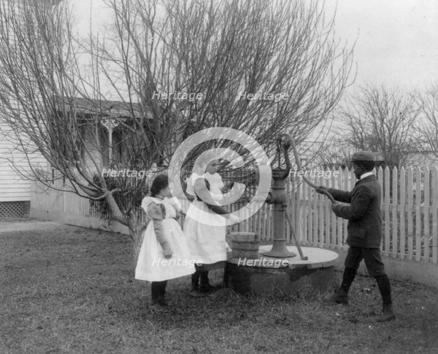 Two girls and a boy pumping water at well of Hampton Institute graduate, 1899 or 1900. Creator: Frances Benjamin Johnston.