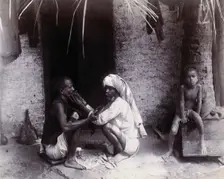 Two Indian men crouching; one is cutting the other's hair, while a girl sits.., c1890s. Creator: Unknown