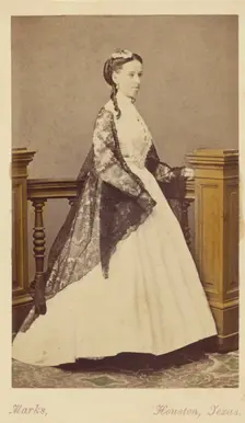 Unidentified woman wearing a white dress and black shawl, standing, 1870-1875. Creator: H. R. Marks