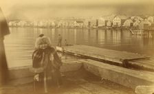 Unidentified young girl standing on wharf, with buildings along waterfront...,1894 or 1895. Creator: Alfred Lee Broadbent