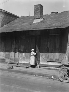 View from across street of a woman with a broom standing in a doorway, New Orleans, c1920-c1926. Creator: Arnold Genthe