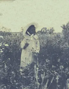 Young African-American(?) girl, wearing long dress and bonnet, standing in field holding..., c1900. Creator: Unknown