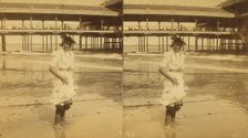 Young woman wading at the beach, in front of a covered pier, (1868-1900?). Creator: Unknown