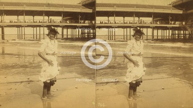 Young woman wading at the beach, in front of a covered pier, (1868-1900?). Creator: Unknown.